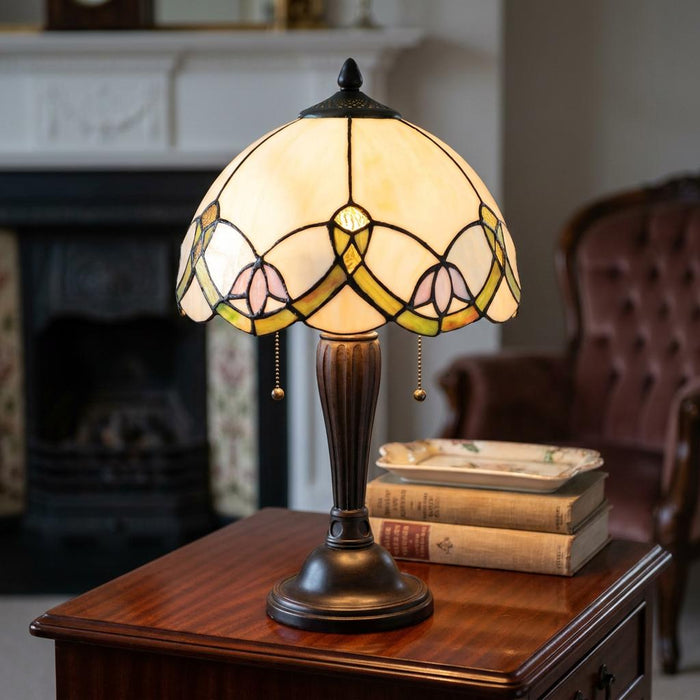 Abbey Tiffany table lamp with a floral cream glass shade and bronze base on a wooden side table with books.