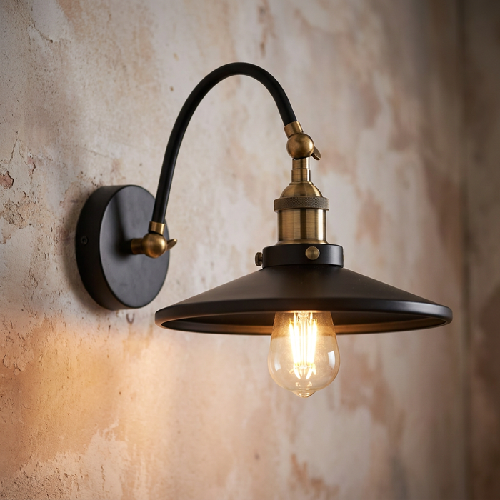 Aken adjustable black and gold wall light with a metal shade and glowing bulb on a rustic wall.