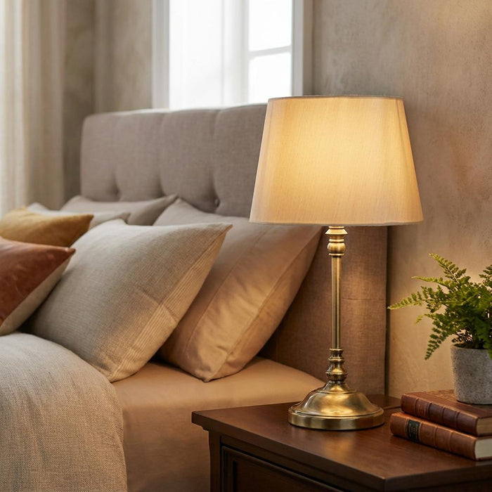 Antique brass bedside lamp with a beige fabric shade on a wooden nightstand in a cozy bedroom setting.
