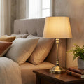 Antique brass bedside lamp with a beige fabric shade on a wooden nightstand in a cozy bedroom setting.