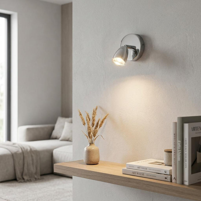 Saxby Amalfi chrome single spotlight on a wall, illuminating a wooden shelf in a modern neutral living room.