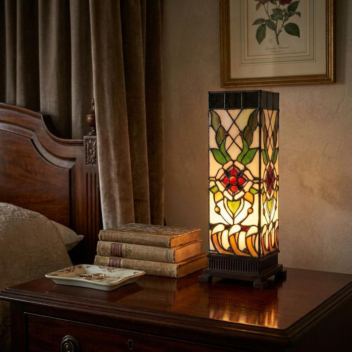 A pillar-shaped floral Tiffany lamp glows on a dark wooden nightstand beside a stack of vintage books.
