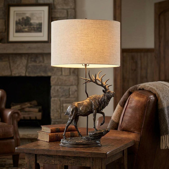 Bronze stag table lamp with a natural fabric oval shade on a wooden side table in a rustic living room setting.