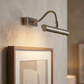 Antique chrome picture light with a curved arm illuminating a framed artwork on a neutral wall.