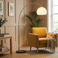 Chrome arc floor lamp with a glowing white dome shade positioned over a mustard yellow armchair in a cozy living room.