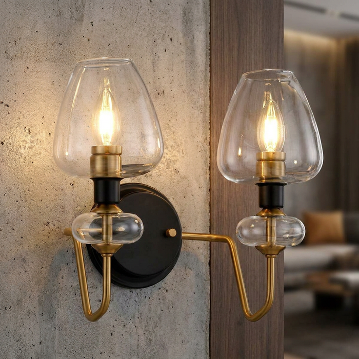 Aged brass wall sconce with two candle bulbs and clear glass shades mounted on a textured concrete wall.