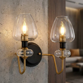 Aged brass wall sconce with two candle bulbs and clear glass shades mounted on a textured concrete wall.