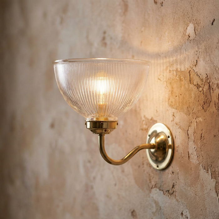 Art Deco wall light with a fluted glass bowl shade and brass arm, glowing against a rustic plaster wall.
