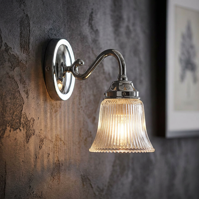 Polished chrome wall sconce with a ribbed glass shade, lit on a dark textured wall.