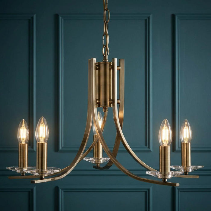 Ascona 5-light antique brass chandelier with glass sconces and lit bulbs against a teal paneled wall.