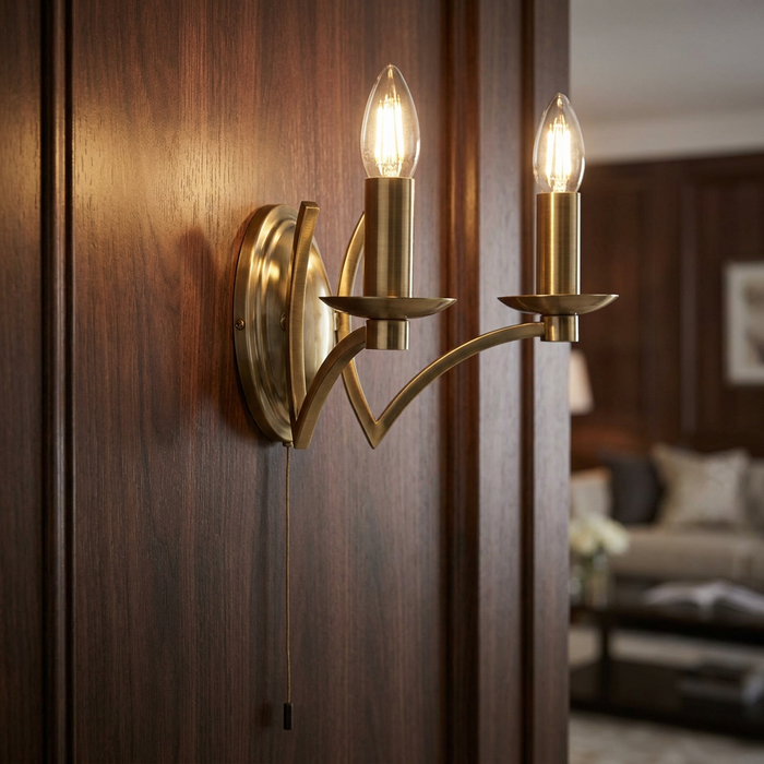 Antique brass wall light with two glowing candle bulbs and a pull cord mounted on a dark wood-paneled wall.