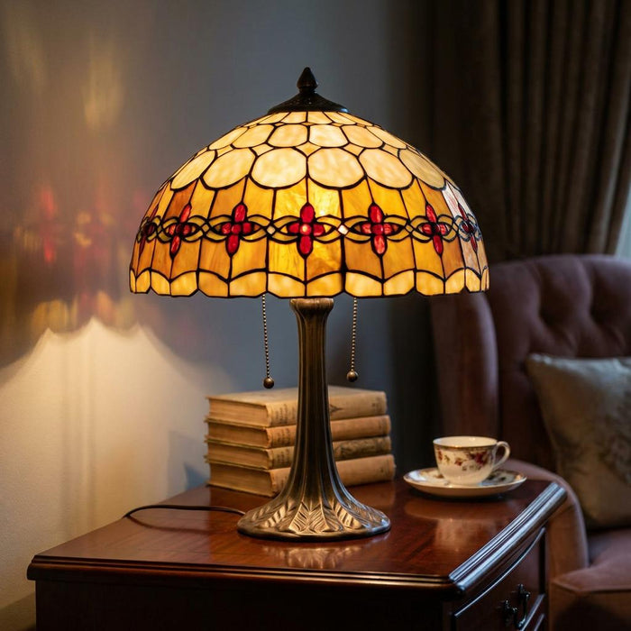 Illuminated Tiffany table lamp with an amber and red stained glass shade on a wooden side table with books.