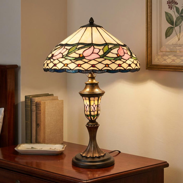 Tiffany table lamp with a pink floral stained glass shade on a dark wood bedside table next to vintage books.