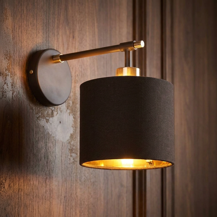 Brown and polished brass wall light with a dark fabric drum shade, mounted on a dark wood-paneled wall.