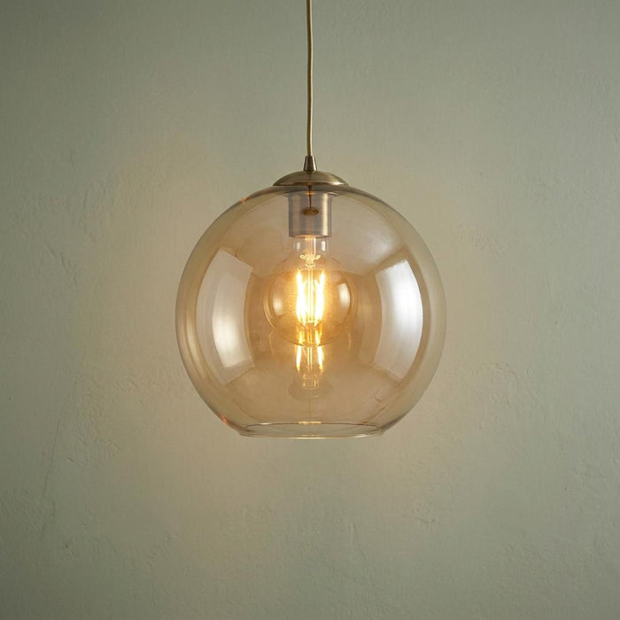Lit 25cm round amber glass ceiling pendant with antique brass fitting against a light textured wall.
