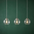 Three clear glass globe pendant lights with glowing Edison bulbs hanging against a dark green textured wall.