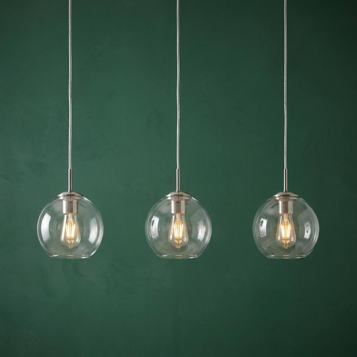 Three clear glass globe pendant lights with glowing Edison bulbs hanging against a dark green textured wall.
