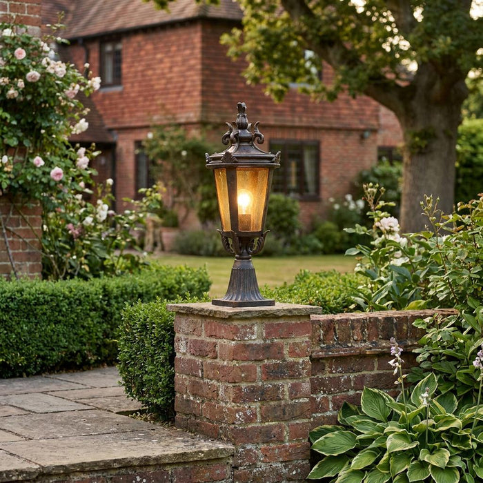 Ornate bronze pedestal lantern lit on a brick garden wall with a traditional house in the background.