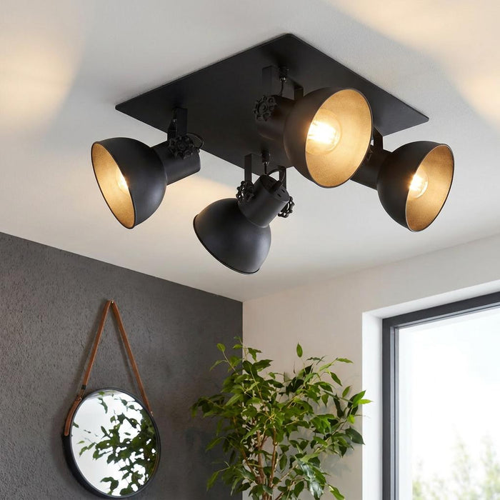 Industrial black 4-light ceiling spotlight on a square plate, illuminating a modern room with a grey wall and plant.