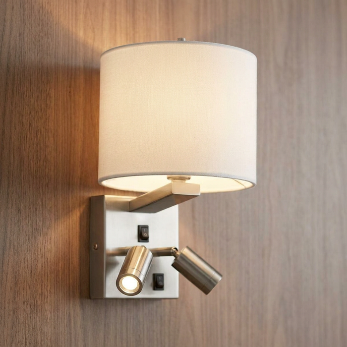 Brushed nickel wall light with a white drum shade and two adjustable spotlights mounted on a wood-paneled wall.