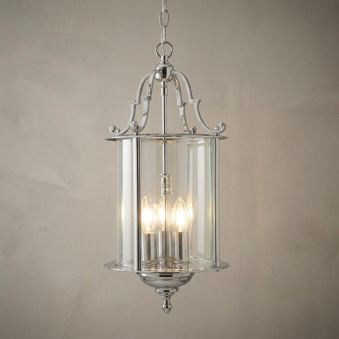 Polished chrome bevelled lantern ceiling pendant with three glowing bulbs against a neutral textured wall.