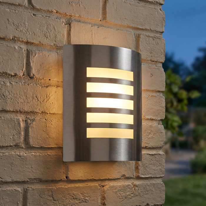 Modern silver outdoor wall light with horizontal slats glowing warmly on a light brick wall at dusk.