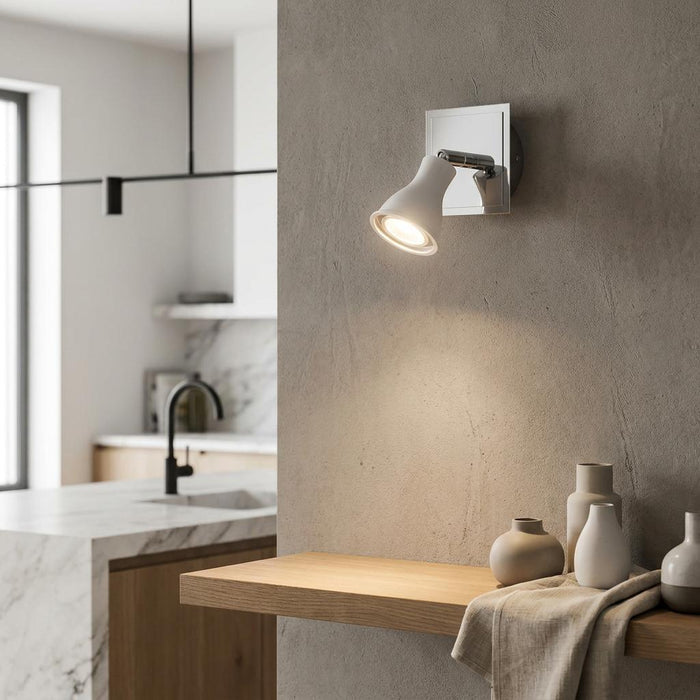 White and chrome single spotlight mounted on a textured wall, illuminating decorative vases on a wooden shelf.