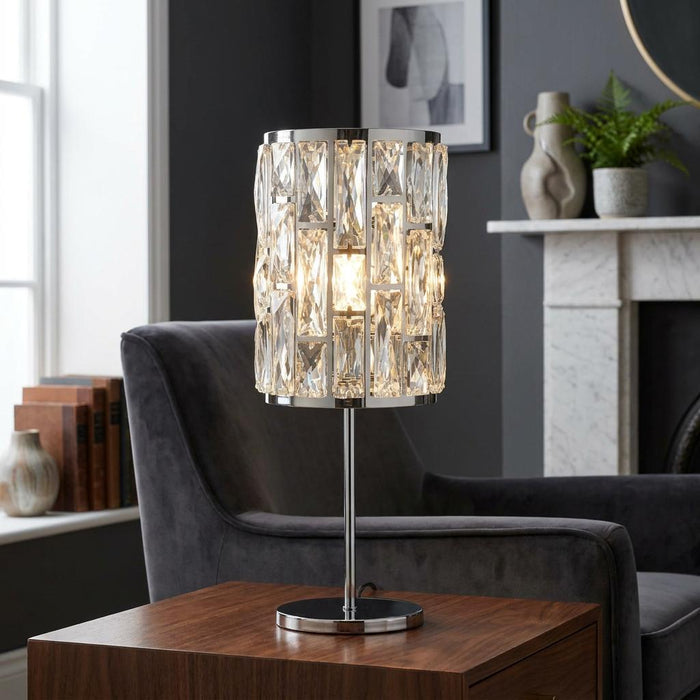 Cylindrical chrome table lamp with crystal glass panels, glowing warmly on a wooden table in a modern living room.