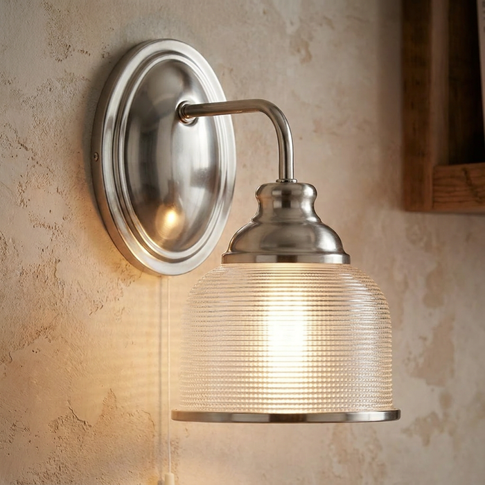 Silver wall light with a ribbed glass shade and pull cord, glowing against a rustic beige wall.