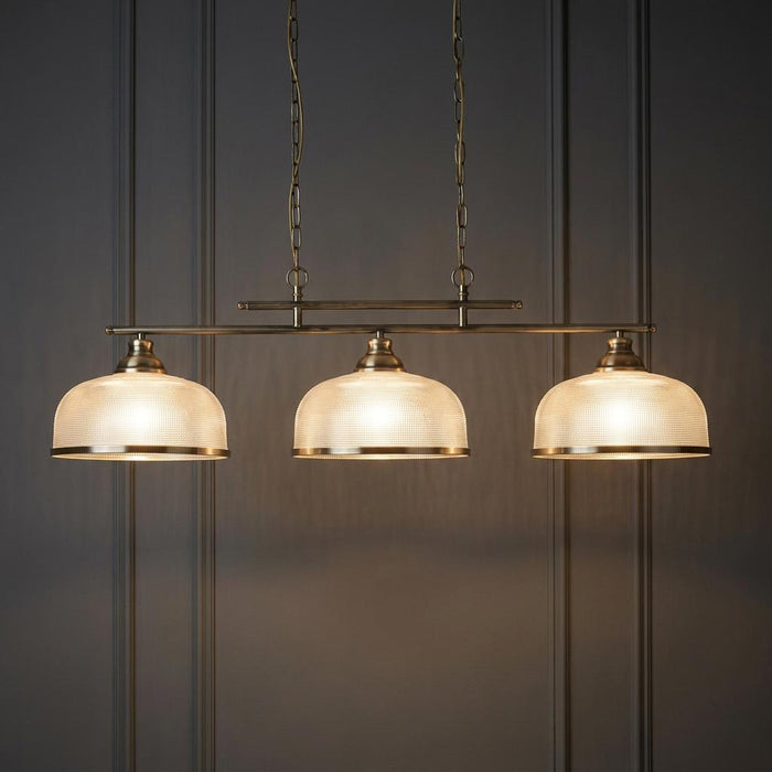Three-light brass bar pendant with holophane glass shades suspended against a dark paneled wall.