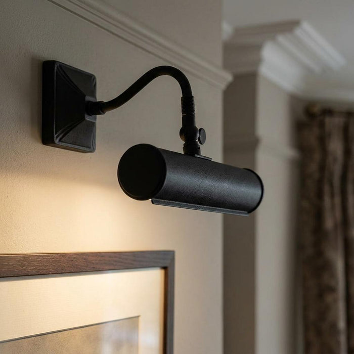 Black wall-mounted picture light with a cylindrical shade illuminating a framed artwork on a cream wall.