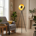 Black tripod floor lamp with a gold leaf interior in a neutral-toned living room beside a grey armchair.