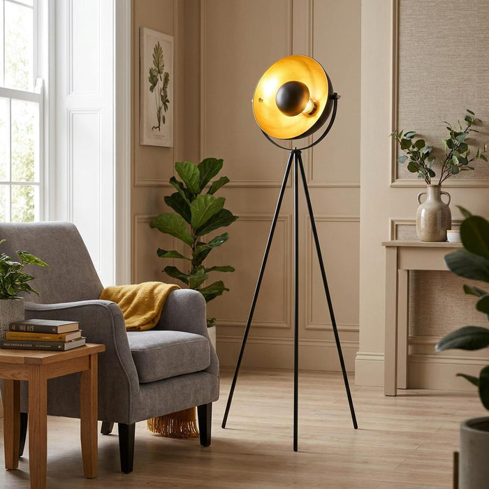 Black tripod floor lamp with a gold leaf interior in a neutral-toned living room beside a grey armchair.