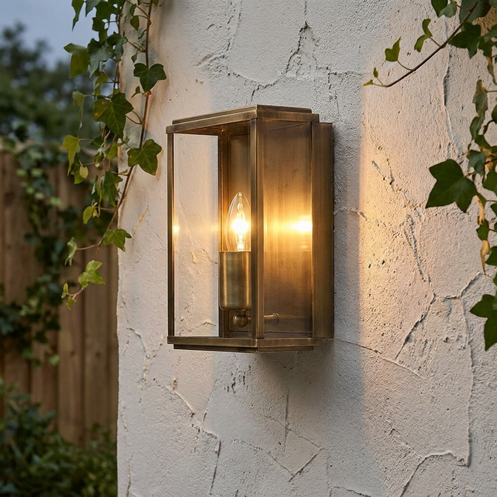 Antique brass box-style outdoor wall lantern lit on a white textured wall with green ivy.