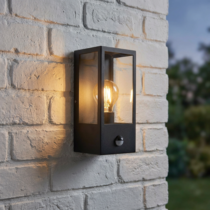 Modern black rectangular lantern with PIR sensor and glowing bulb mounted on a light brick wall.
