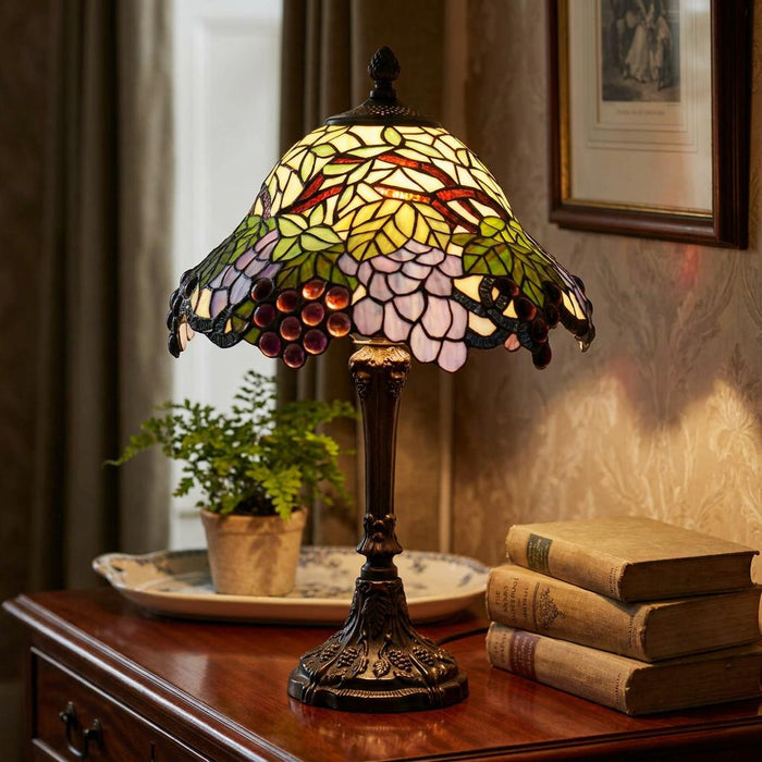 Tiffany table lamp with grape-patterned stained glass shade glowing on a wooden sideboard next to vintage books.