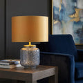 Brondo cobalt mottled glass table lamp with a gold drum shade on a dark wooden table beside a navy velvet armchair.