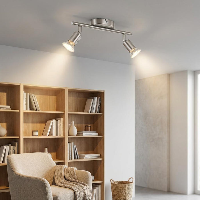 Brushed nickel 2-light bar spotlight mounted on a ceiling, illuminating a bookshelf in a modern room.