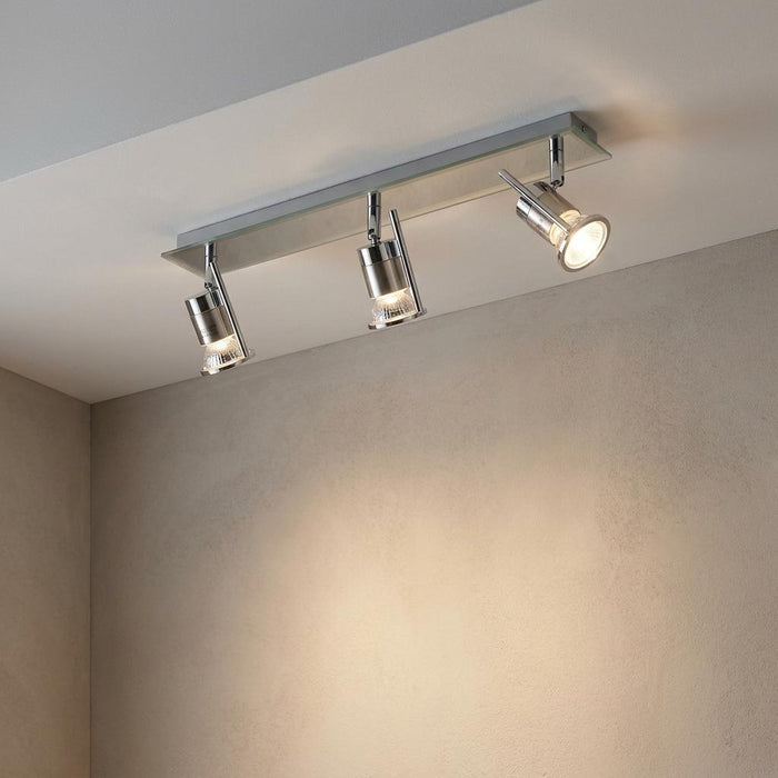 Modern Cara chrome 3-light bathroom ceiling spotlights emitting a warm glow against a minimalist beige wall.