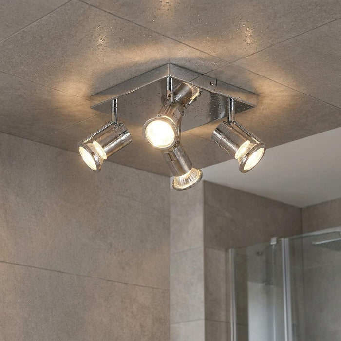 Polished chrome four-light bathroom spotlight fixture illuminated on a grey tiled ceiling.