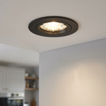 Matt black recessed downlight illuminated in a white ceiling, casting a warm glow on a wall.