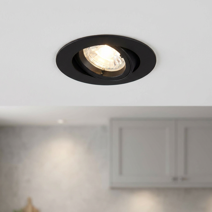 A matt black tilt recessed downlight illuminated on a white ceiling above a blurred kitchen.