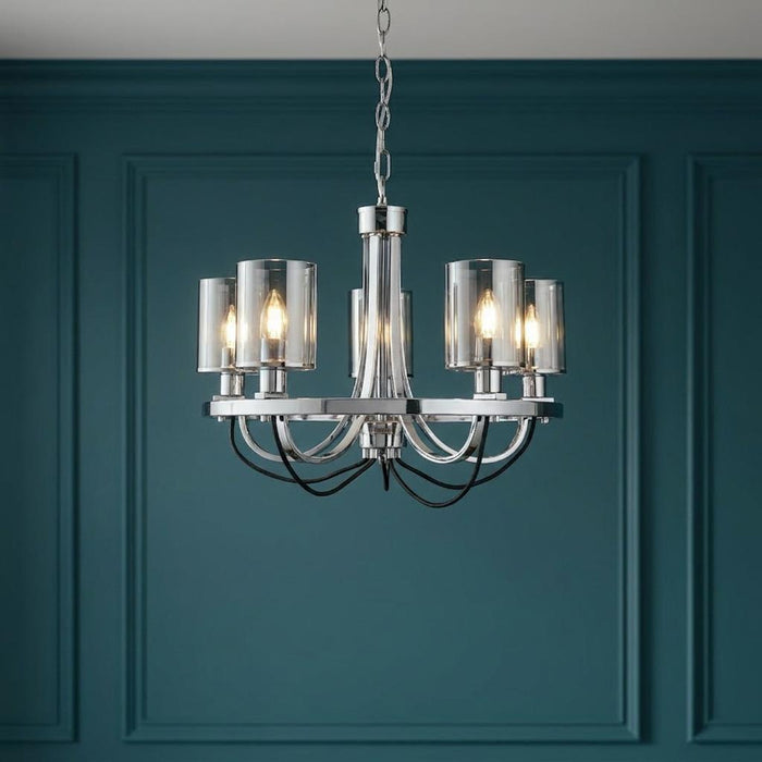 Five-light chrome chandelier with cylindrical smoked glass shades, illuminated against a teal paneled wall.
