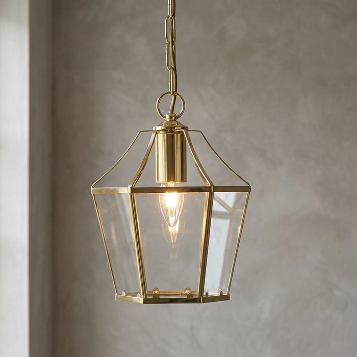 Polished brass lantern pendant with clear glass panels hanging against a textured grey wall.