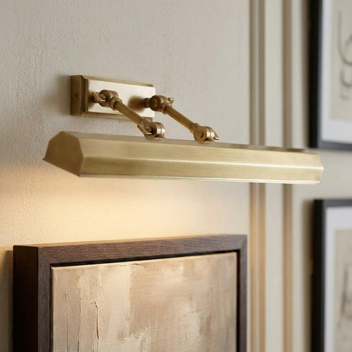 Aged brass picture light with adjustable arms illuminating a framed abstract painting on a neutral wall.