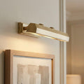 Aged brass picture light mounted on a neutral wall, casting light downward onto a framed abstract painting.