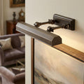 Chawton medium dark bronze picture light with adjustable arms illuminating a landscape painting in a living room.