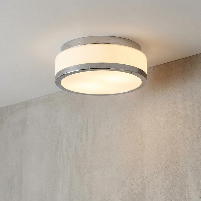 Modern chrome-rimmed flush ceiling light with an illuminated white glass shade against a textured grey wall.