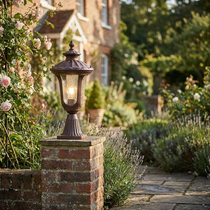 Ornate bronze pedestal lantern lit on a brick pillar along a cottage garden path with lavender and roses.