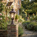 Ornate bronze pedestal lantern lit on a brick pillar along a cottage garden path with lavender and roses.
